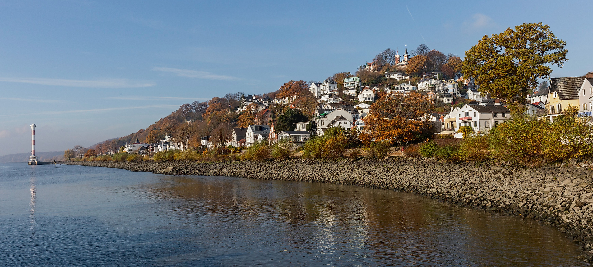 Weiße Häuser am Elbufer in Blankenese vor herbstlich gefärbtem Hang unter blauem Himmel
